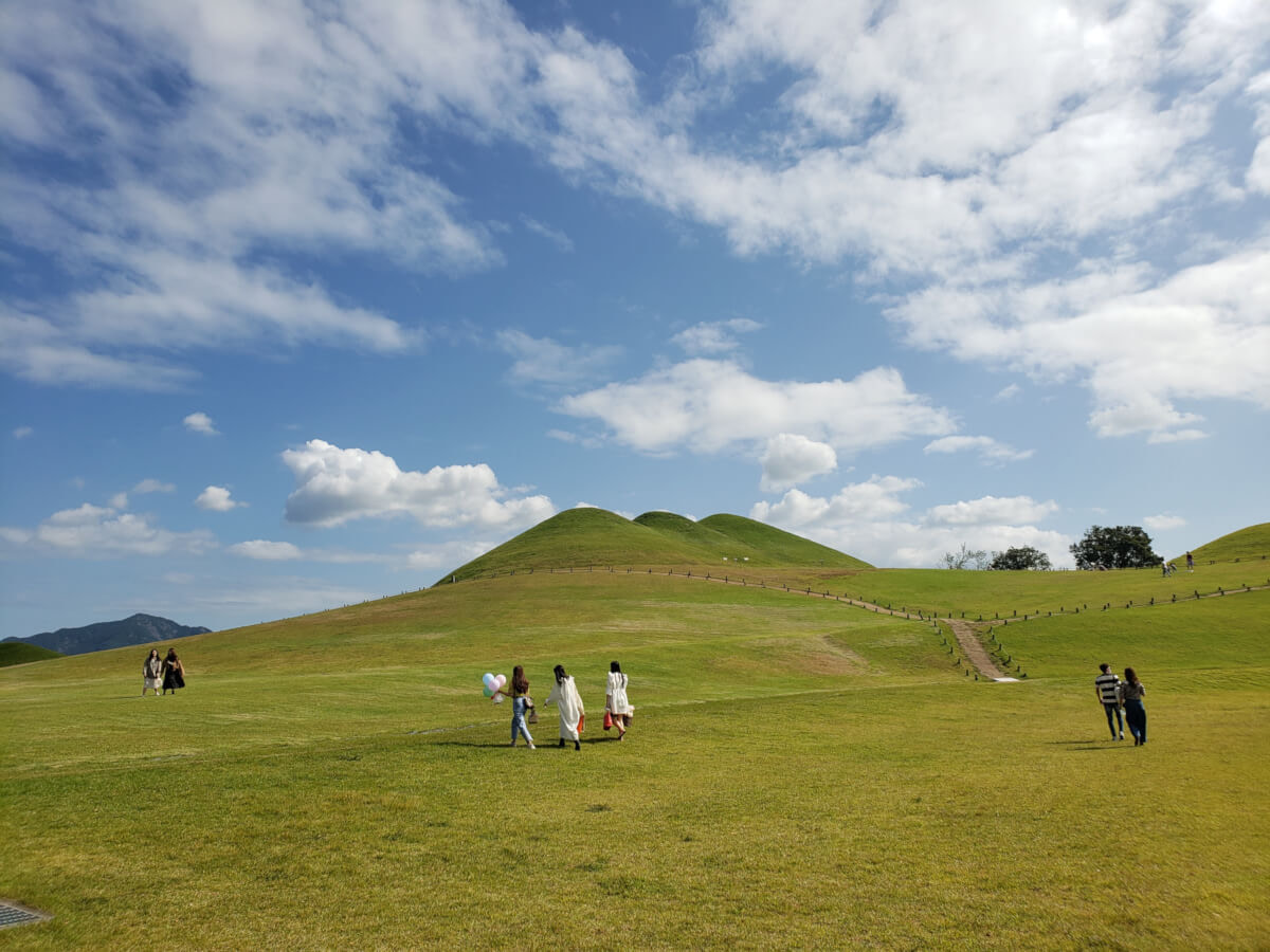 People walking around grassy Songhakdong tomb site on a sunny day with beautiful clouds