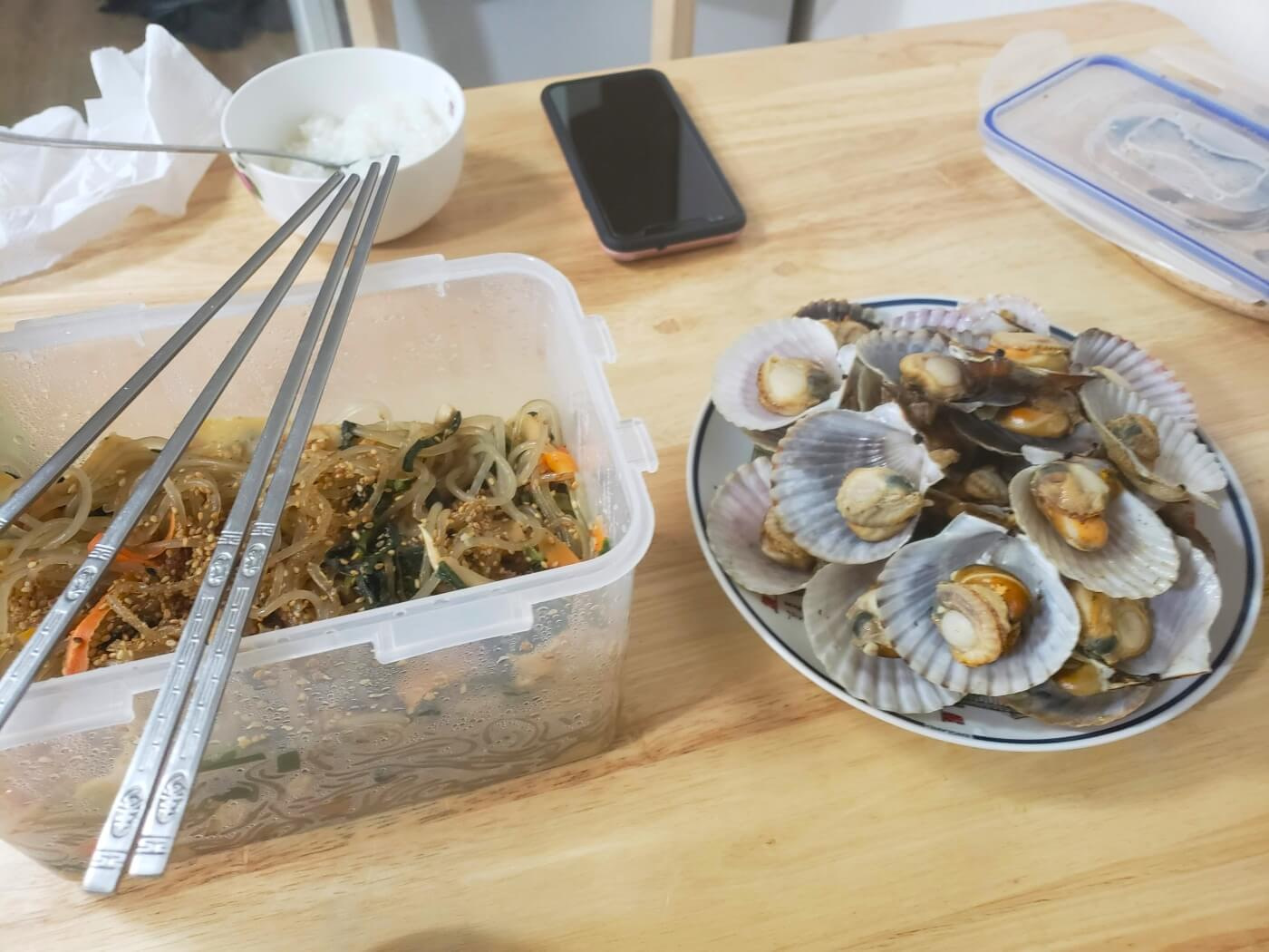 Home-made japchae noodles in plastic tupperware next to a plate of shells