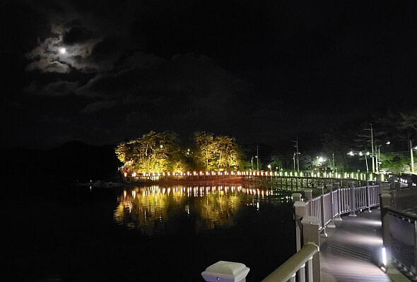 A boardwalk near Haejigae Bridge with the moon shining above lit up trees at night