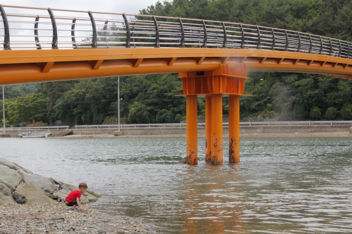 A toddler squatting in the water while playing under an orange bridge.