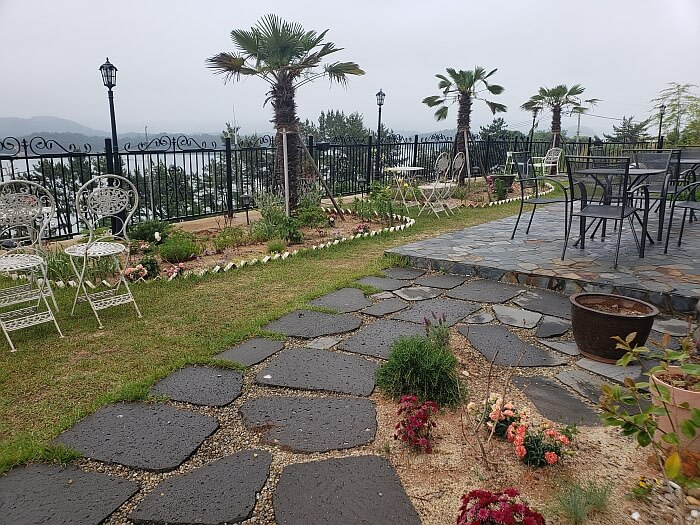 A stone pathway through a courtyard with tables and chairs along a railing and palm trees and flowers with a nice view