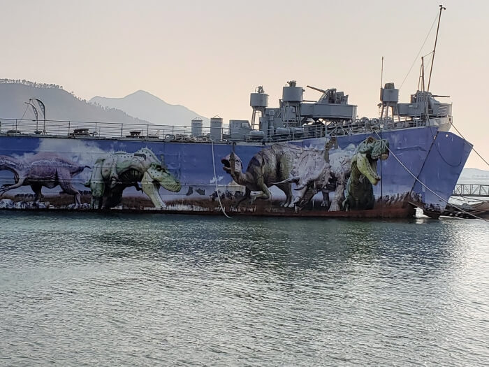 A closeup of a large naval battleship decorated with dinosaur graphics sitting on the water in front of a background of mountains in the setting sun