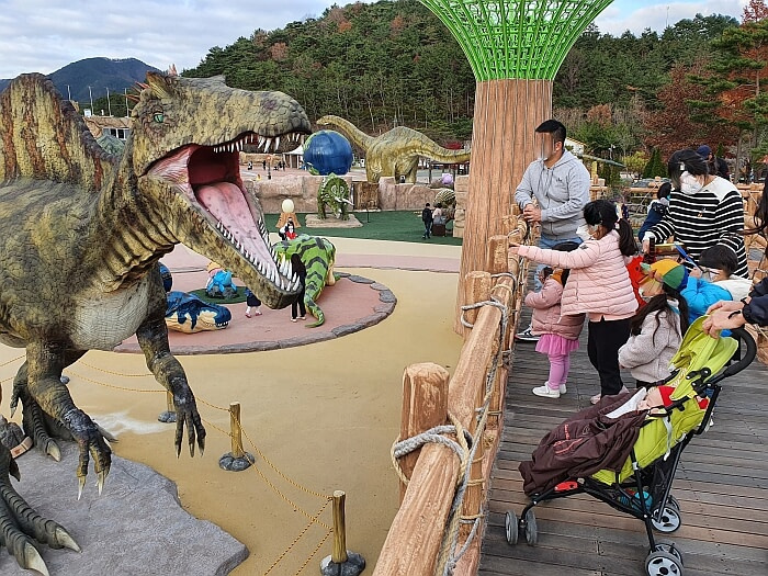 Kids looking at a roaring dinosaur on a platform with various dinosaur characters below and a mountainous background