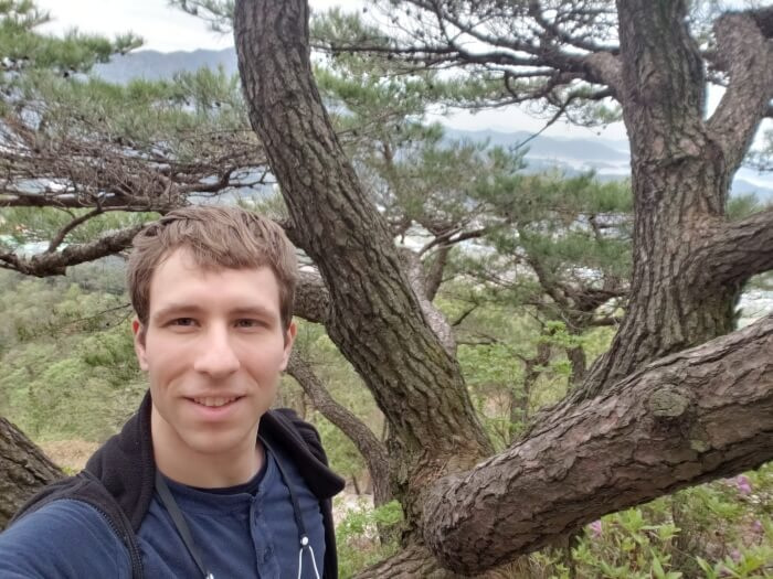 Nate standing next to the tree with winding trunks and branches facing with a nice view in the background