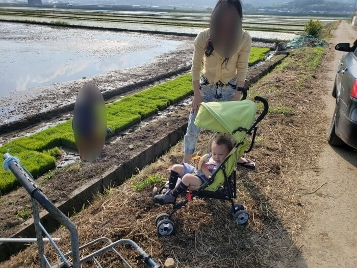 Baby Noah in the stroller with a banana watching my mother-in-law in the field and my wife standing next to the stroller