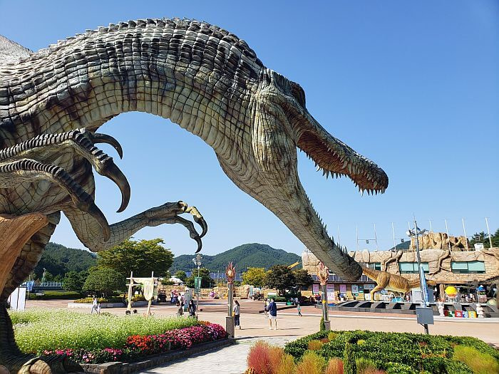 A dinosaur with short arms and sharp claws with an open mouth near a garden and flowering plants