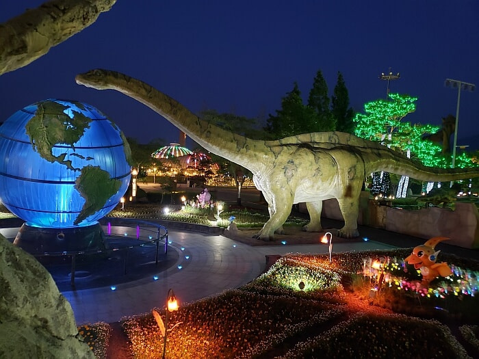 A long-necked dinosaur looming over a globe lit up at night next to gardens and play dinosaurs