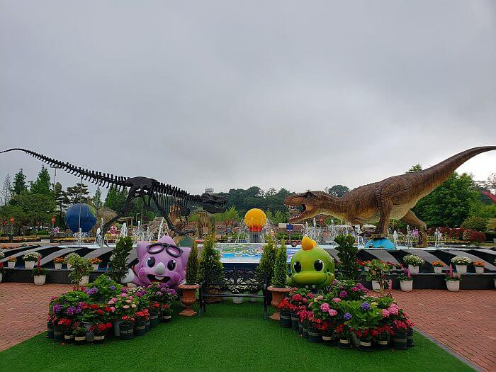 A pair of t-rex dinosaurs in a fountain with the one on the left being a bronze skeleton and the one on the right with flesh and a photo area in front of it with flowers and small play dinosaurs