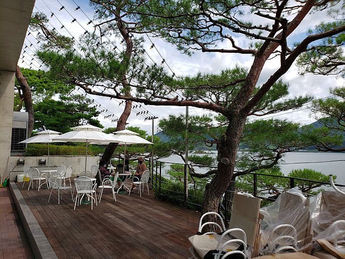 An outdoor deck with umbrellas over tables and chairs with trees mixed in and a view of the sea