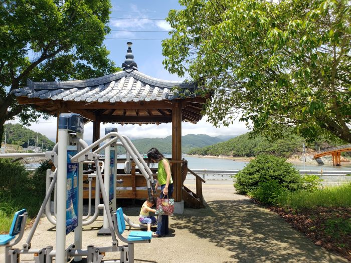 A mother and son at a gazebo next to exercise equipment on a summer day near an island in Korea.