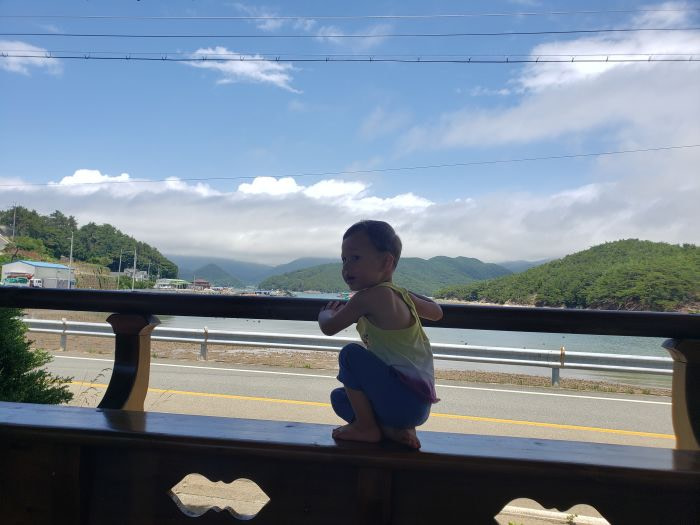 A toddler sitting in a wooden gazebo in Korea overlooking an island in the sea.