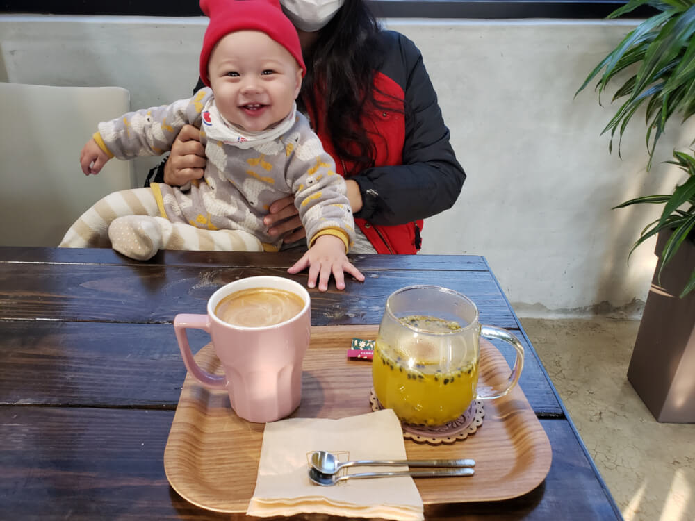 Alice holding Noah who is smiling with a red hat sitting across from a tray of passion fruit tea and latte