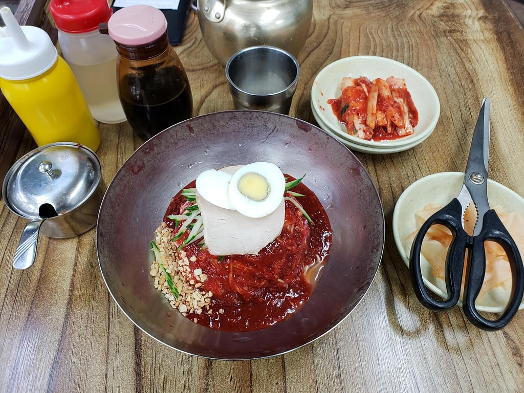 A bowl of spicy milmyeon with raw fish, egg, sauce, and shredded cucumber