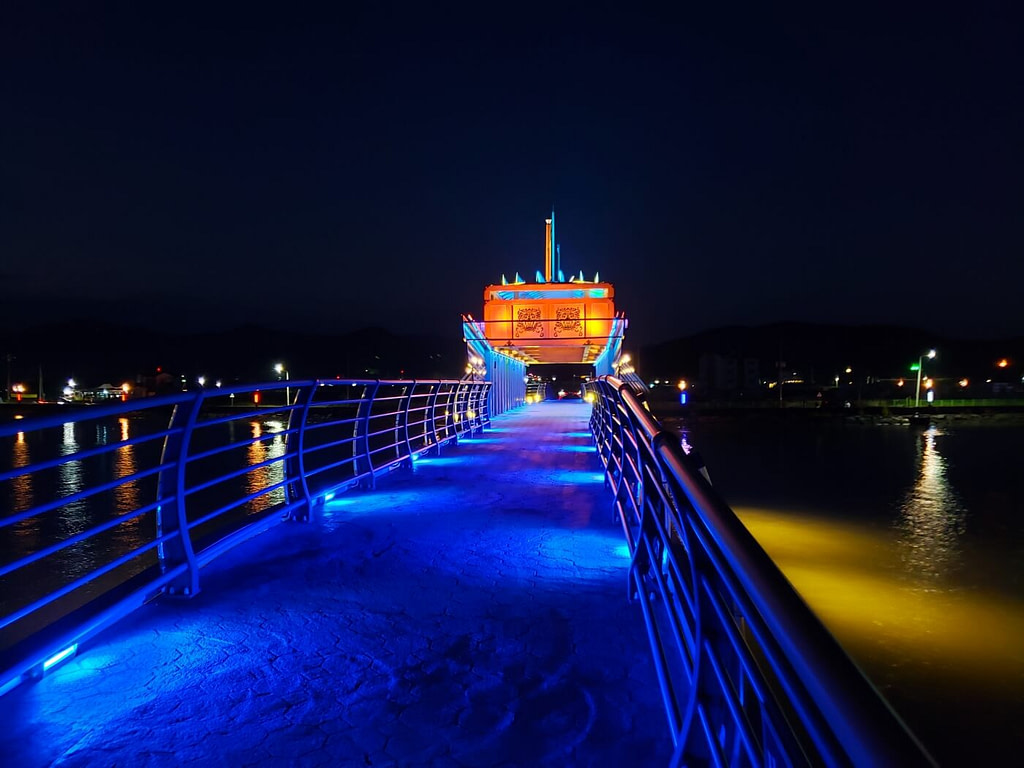 A pedestrian bridge lit up in blue with a turtle ship just ahead over the bridge