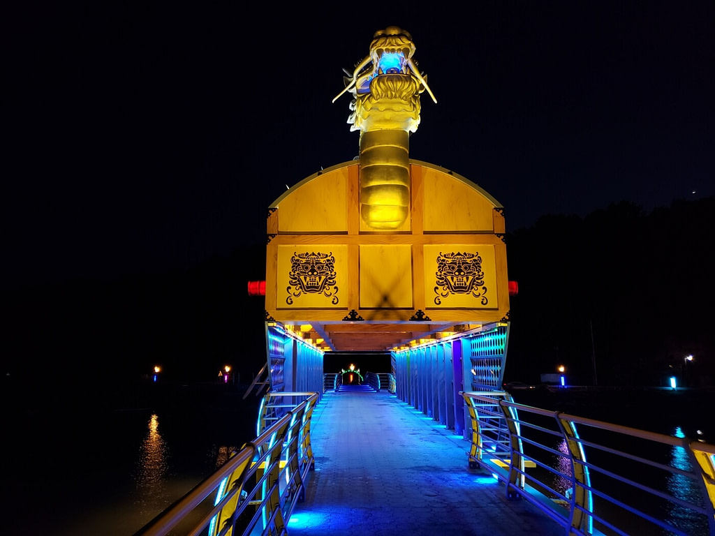 Colorful lights illuminating a turtle ship at night over a walking bridge