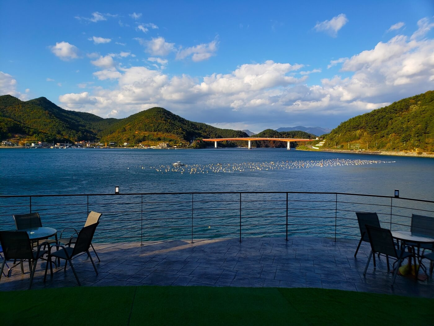 Tables and chairs along a fence by the water with white clouds and blue sky