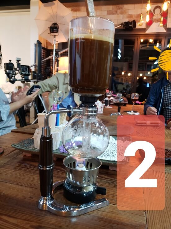 A siphon filled with coffee grounds while the boiling water in a lower glass chamber begins to rise through the siphon