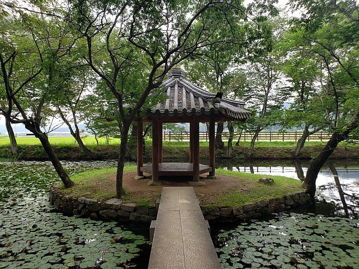 A narrow cement bridge connecting to a small island with a gazebo at the center of a pond surrounded by trees