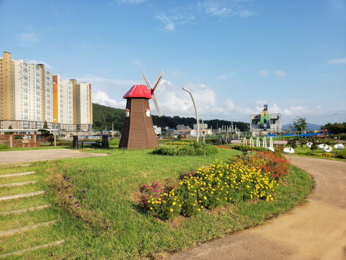 Flowering plants bordering the small windmill on the hill with apartments in the background against a blue sky