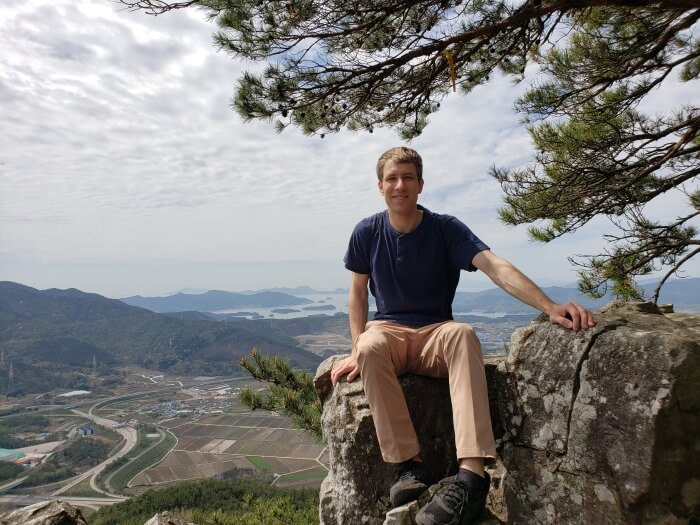 Nate sitting on a rock next to a pine tree climbing Mt. Georyusan with a background of islands in the sea and towns below