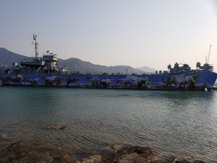 A large naval battleship decorated with dinosaur graphics sitting on the water in front of a background of mountains in the setting sun