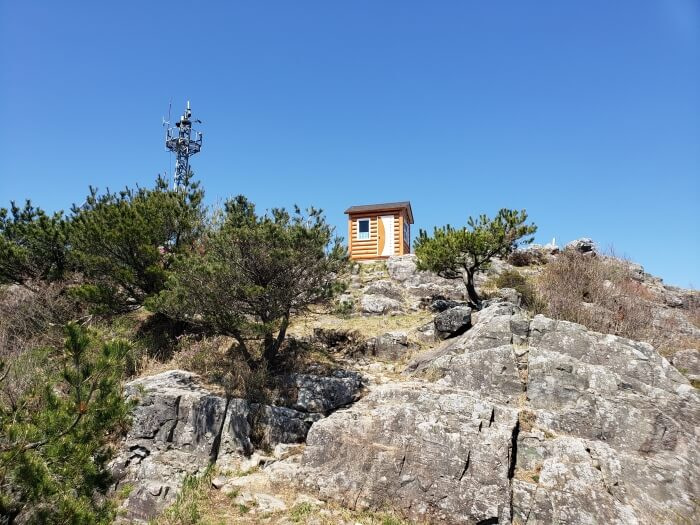 The little cell or radio tower next to the small shelter on the peak with a bunch of large rocks in the foreground