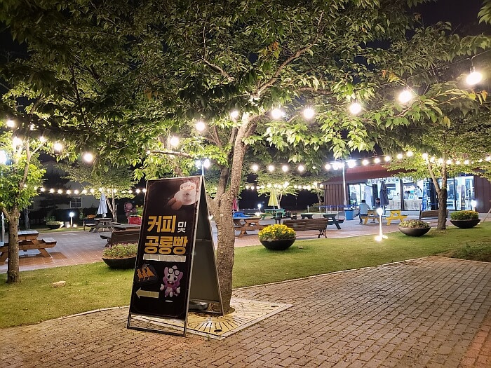 A sign outside under some trees with stringed lights at night in front of a café
