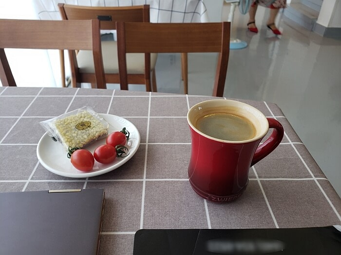 A cup of coffee in a red mug on a table with a plate of a rice snack and cherry tomatoes