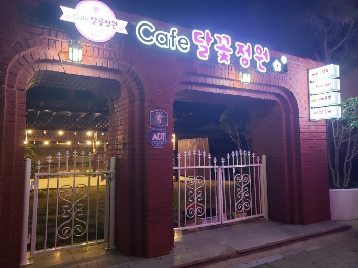 A purple and white sign for Dalgotjeongwon Café lit up at night over an iron gate and brick archway