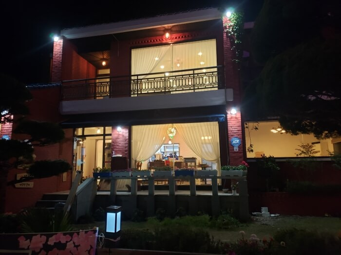 A front view of Dalgotjeongwon Café at night facing the front window with lights illuminating the interior from outside and trees and plants bordering the building