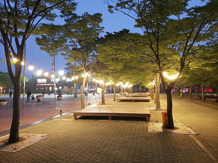 A row of wooden platforms with string lights hung along two rows of trees in the late evening