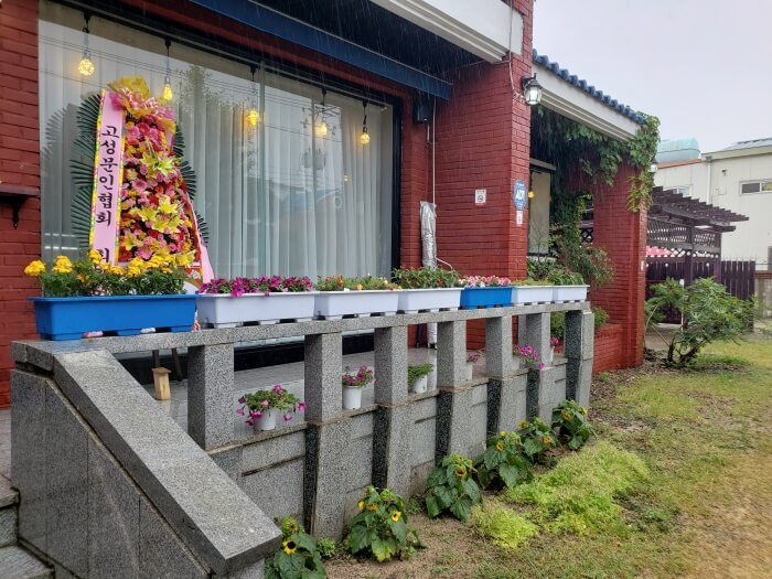 Flowers arranged in trays outside on the front porch along a large window with a white curtain and lights