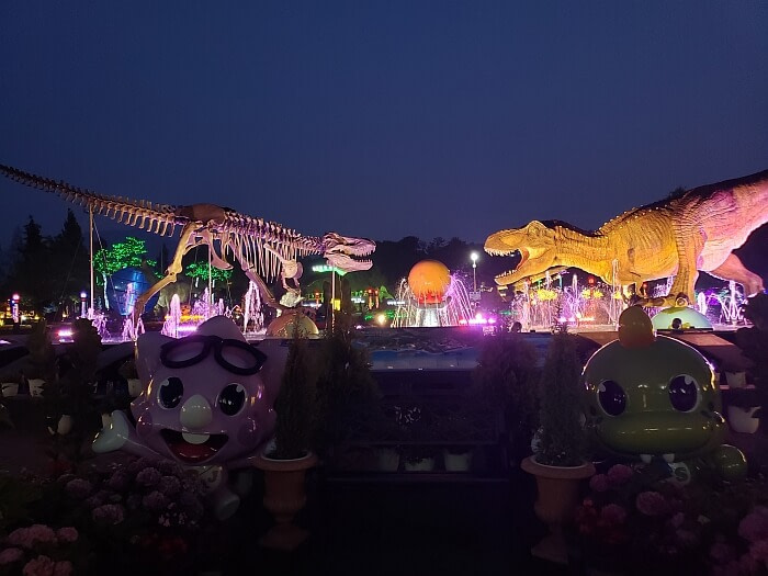 Two dinosaurs lit up at night in a fountain with one dinosaur a skeleton and the other with flesh