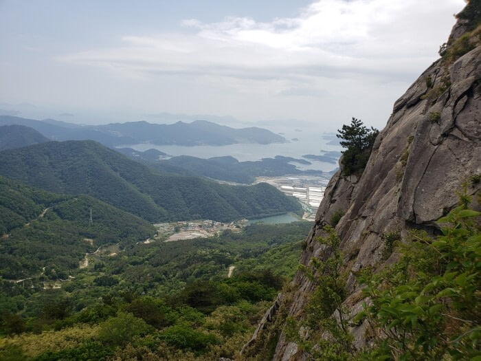 The side of a steep, rocky cliff on Byeogbangsan against a beautiful view of green peaks below and islands in the sea