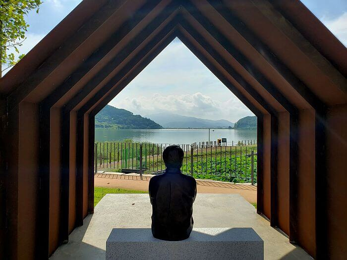 Je Jong-gu statue seated facing water underneath a pointed steel structure of ironclad ships