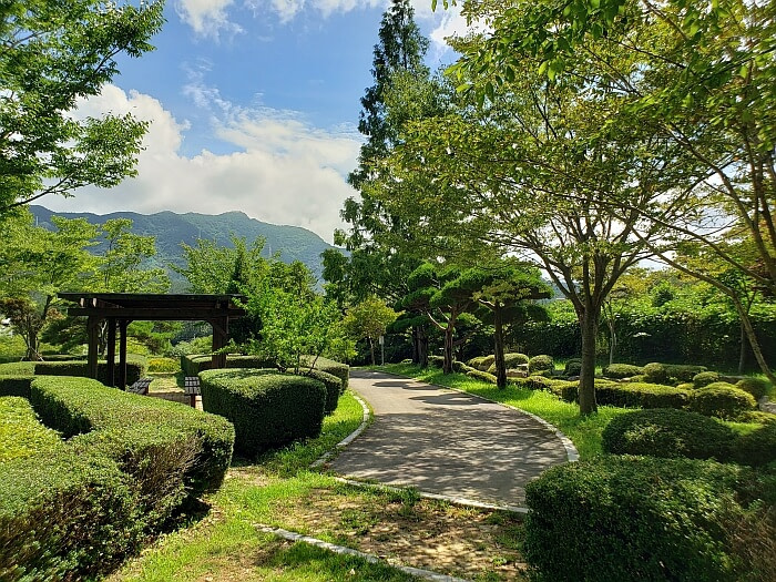 A paved path between bushes with a covered resting area with benches on the side