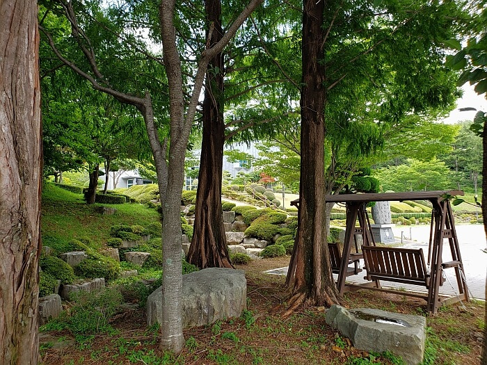 A swinging bench among trees and rocks next to the parking lot