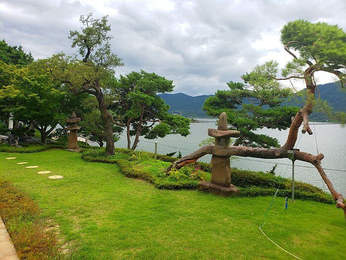 A grassy area with a tree growing sideways and some other trees and and stone statue at the edge of a cliff with a view of the sea and mountains