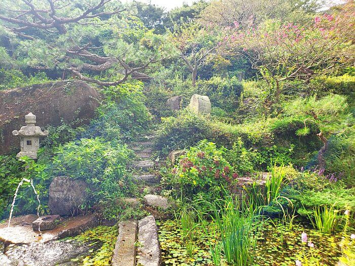 A small, stone bridge across a small pond in a lush garden up a hill with trees and bushes
