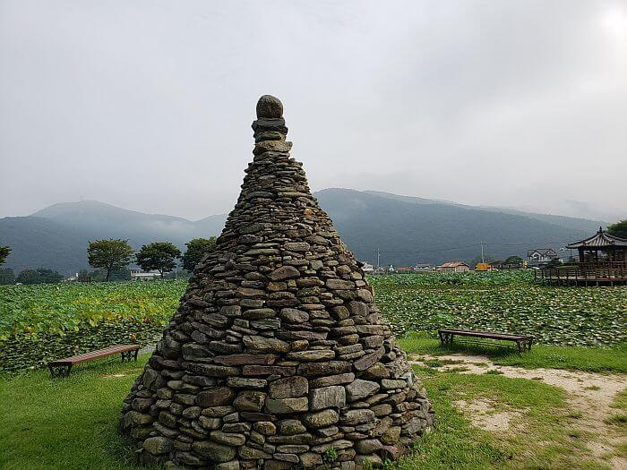 A stone pagoda in the grass surrounded by a bench and the pond