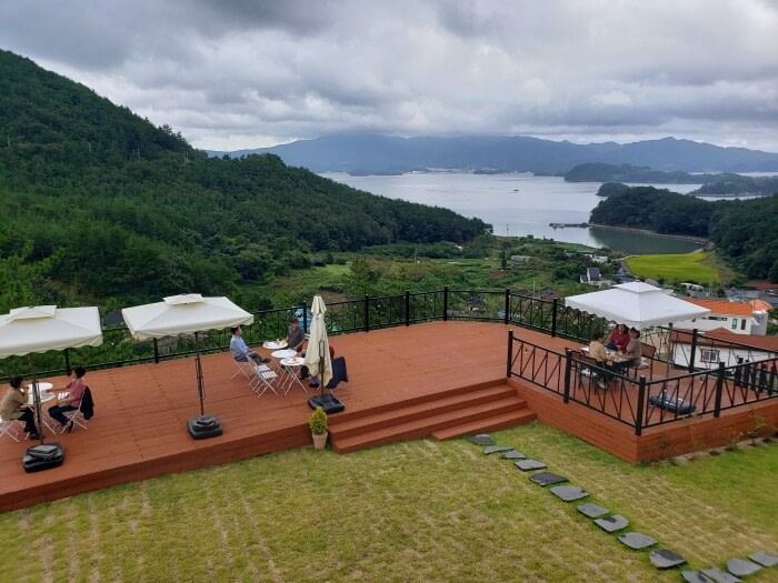 People sitting at tables covered by umbrellas on an outdoor patio overlooking a view of the sea and mountains