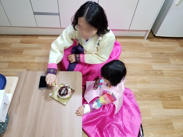 A woman and a little girl both dressed in traditional Korean clothes and sitting on the floor at a small table with a small cake