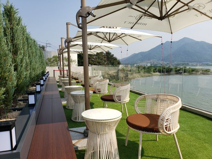 Seating on a rooftop lined with small evergreens and umbrellas facing a sea and mountain view