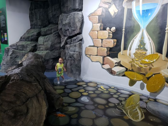 A toddler next to painted trick art with an hourglass and coins falling into water