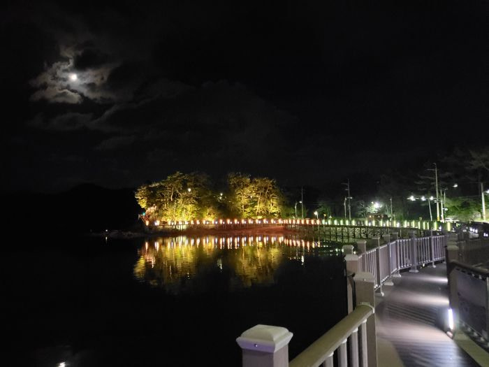A boardwalk near Haejigae Bridge with the moon shining above lit up trees at night