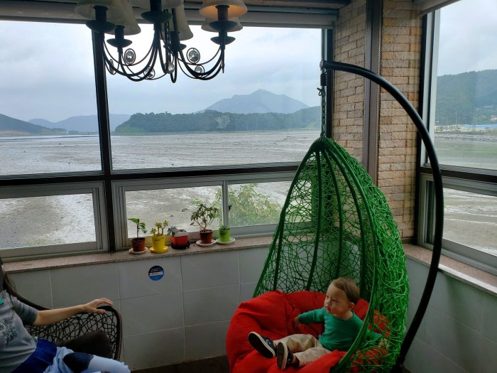 A toddler in a swinging basket in the corner of some windows with a view of the sea